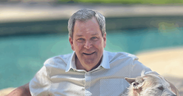 A person in a light shirt is smiling outdoors, accompanied by a goat. There's water in the background.