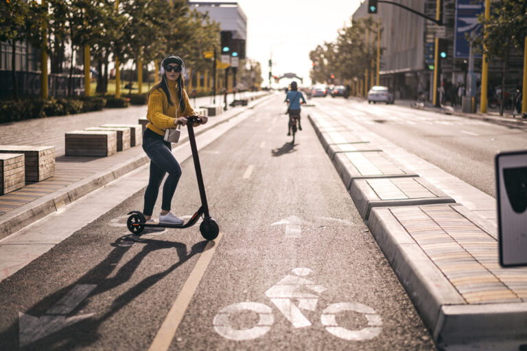 A person is riding an electric scooter on a sunny street, while another rides a bicycle in the background.